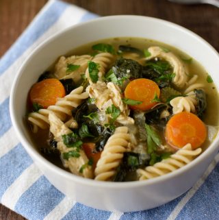 A bowl filled with shredded chicken, kale, noodles, sliced carrots and chicken broth.