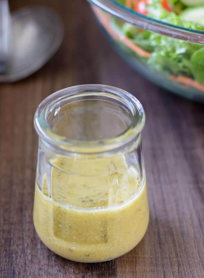 A glass jar with apple cider vinaigrette dressing and a large green salad in the background. 